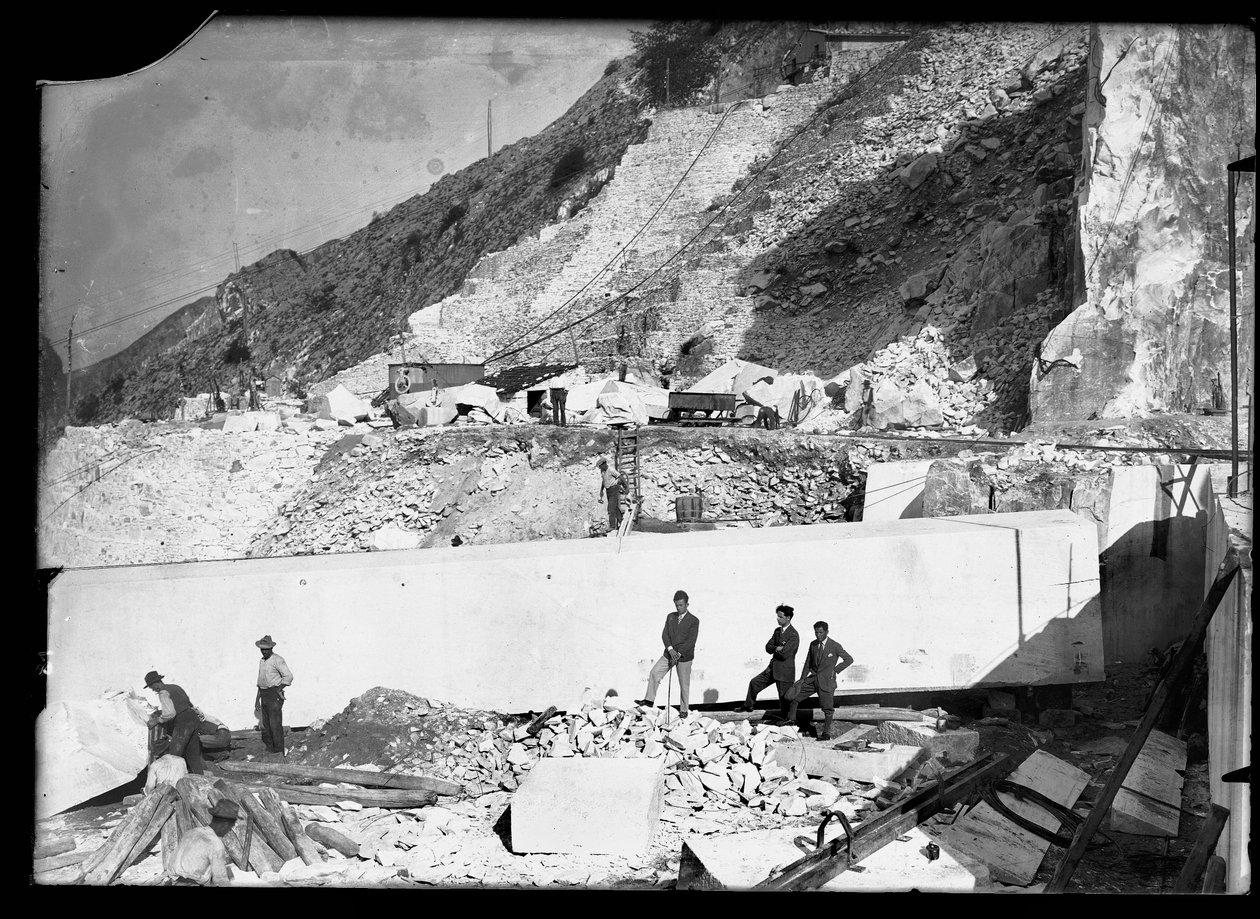 Tre uomini osservano il lavoro di diversi escavatori nelle Alpi Apuane. Dietro di loro un enorme blocco di marmo già squadrato e pronto per essere trasportato a valle da A. Corsini