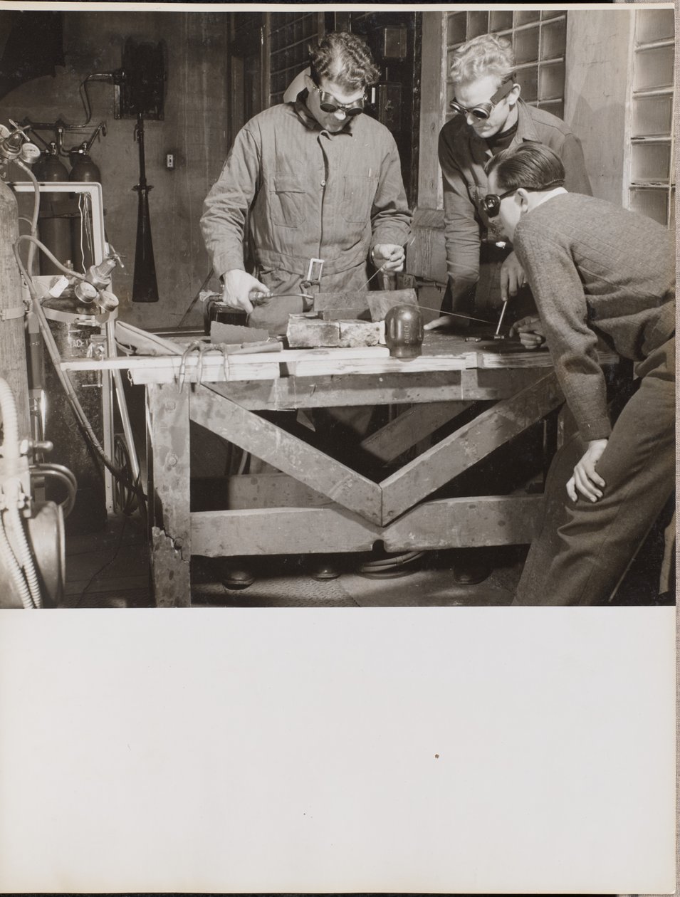 [Tre uomini con gli occhiali saldano un pezzo di metallo su un tavolo.] da Arnold Eagle