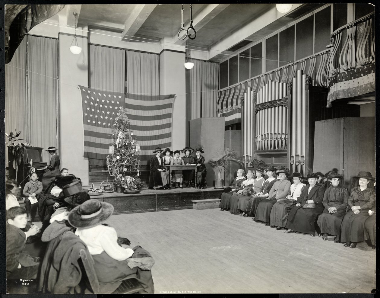 Persone in un auditorium presso la New York Association for the Blind con albero di Natale e persone che forse eseguono una scenetta sul palco con organo, 1920 da Byron Company