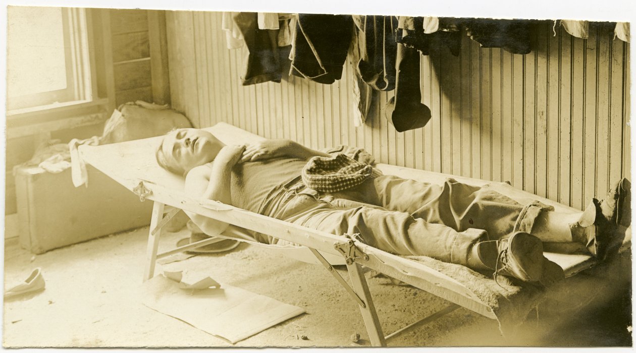 Fotografia orizzontale seppia che mostra un uomo che dorme su una branda in un edificio con pannelli di legno. Vari articoli di abbigliamento possono essere visti appesi al muro e valigie possono essere viste a terra nell