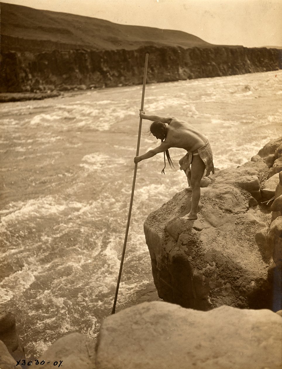 Indiano Tlakluit, in piedi su una roccia, pesca con una rete da immersione, 1910 circa (foto b/n) da Edward Sheriff Curtis