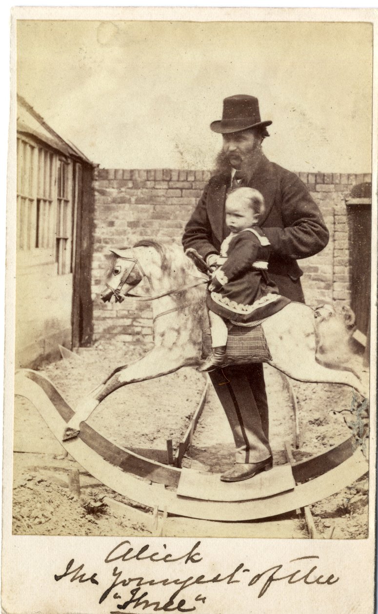 Bambina su un cavallo a dondolo in giardino, con il padre accanto da English Photographer