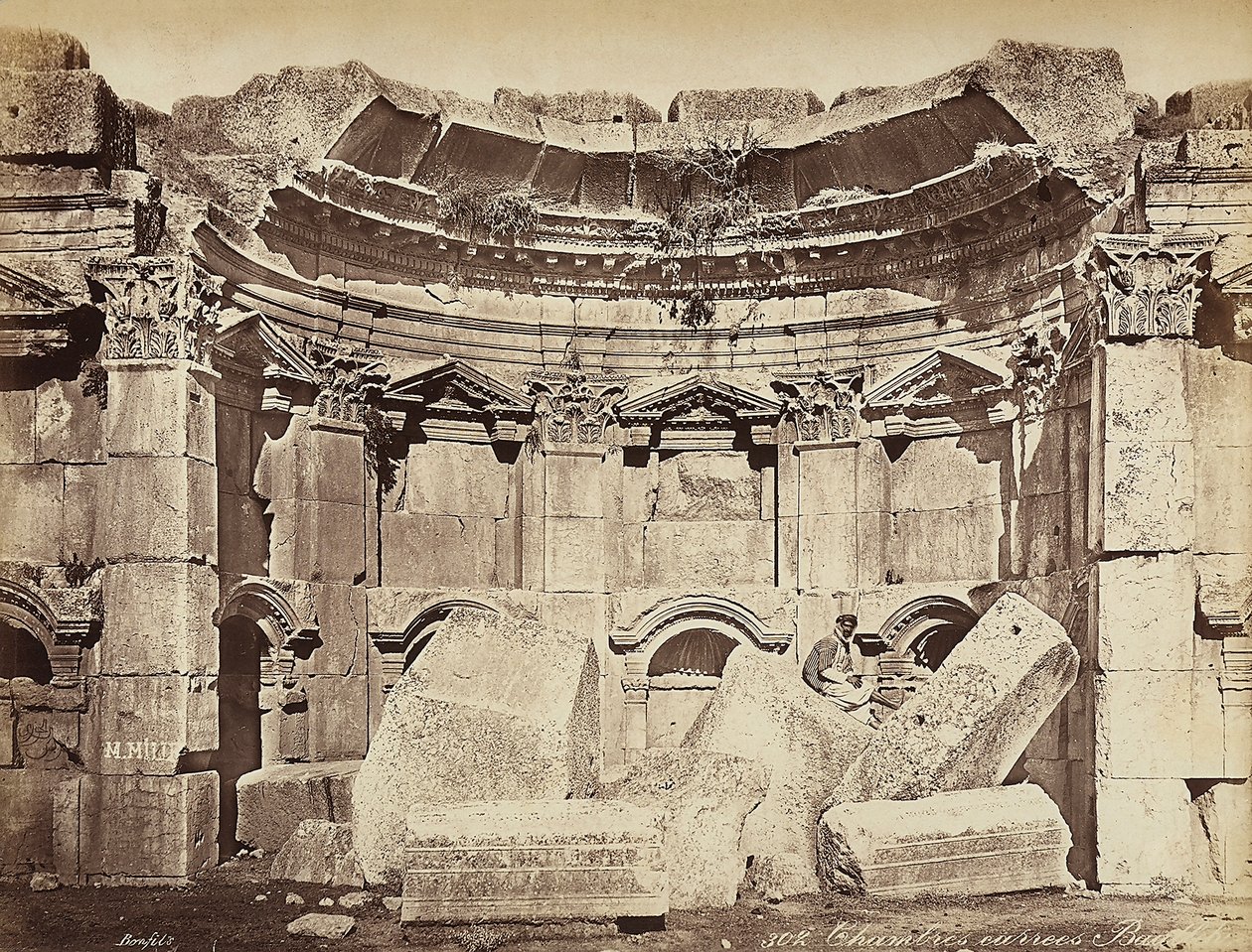 Dettaglio delle cosiddette "stanze" nella zona archeologica di Heliopolis o Baalbek, antica città siriana, ora Libano. Un uomo posa, seduto sulle rovine da Félix Bonfils