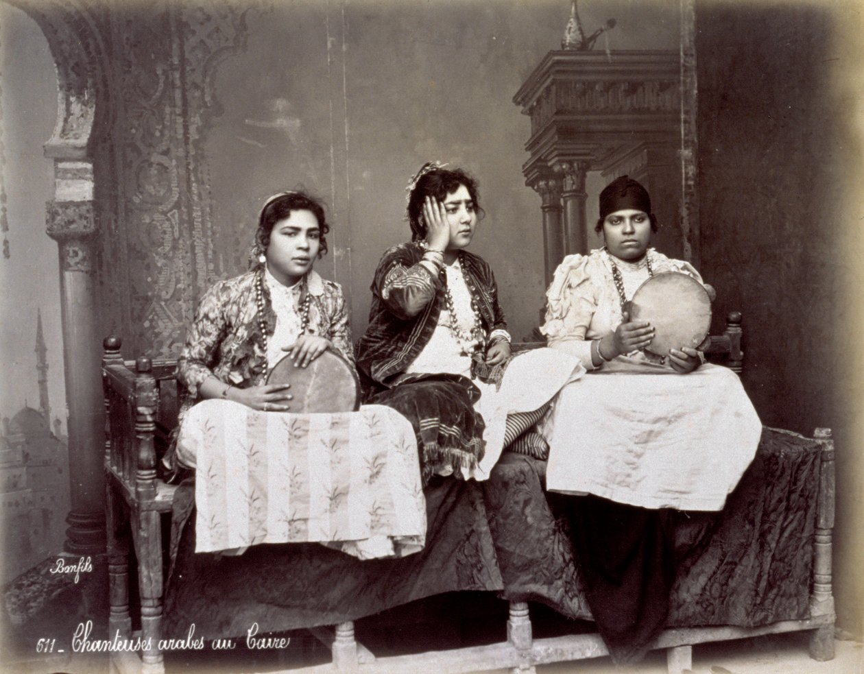 Ritratto di tre giovani cantanti arabi in abiti etnici con lunghe collane. Posano in studio, seduti. Due di loro tengono un tamburello da Félix Bonfils