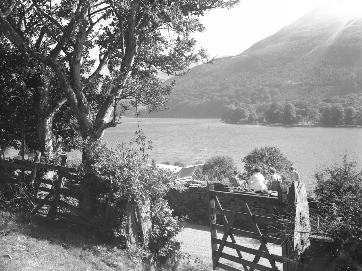 Quattro persone sedute su un muretto a secco a fare un picnic con vista sul lago, auto parcheggiata con il bagagliaio aperto da Joseph Hardman