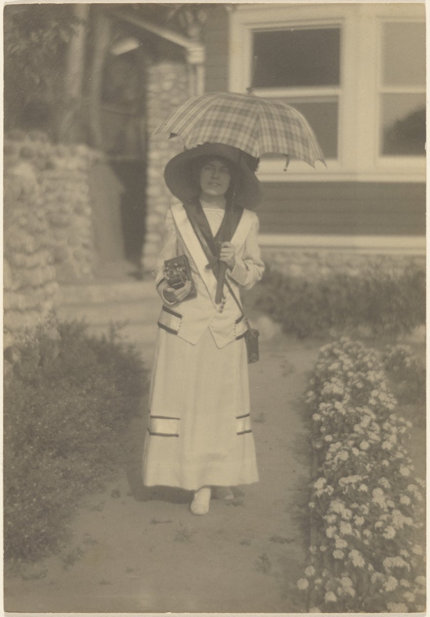 [Donna in piedi nel giardino davanti casa con una macchina fotografica e un ombrello a quadri] da Louis Fleckenstein