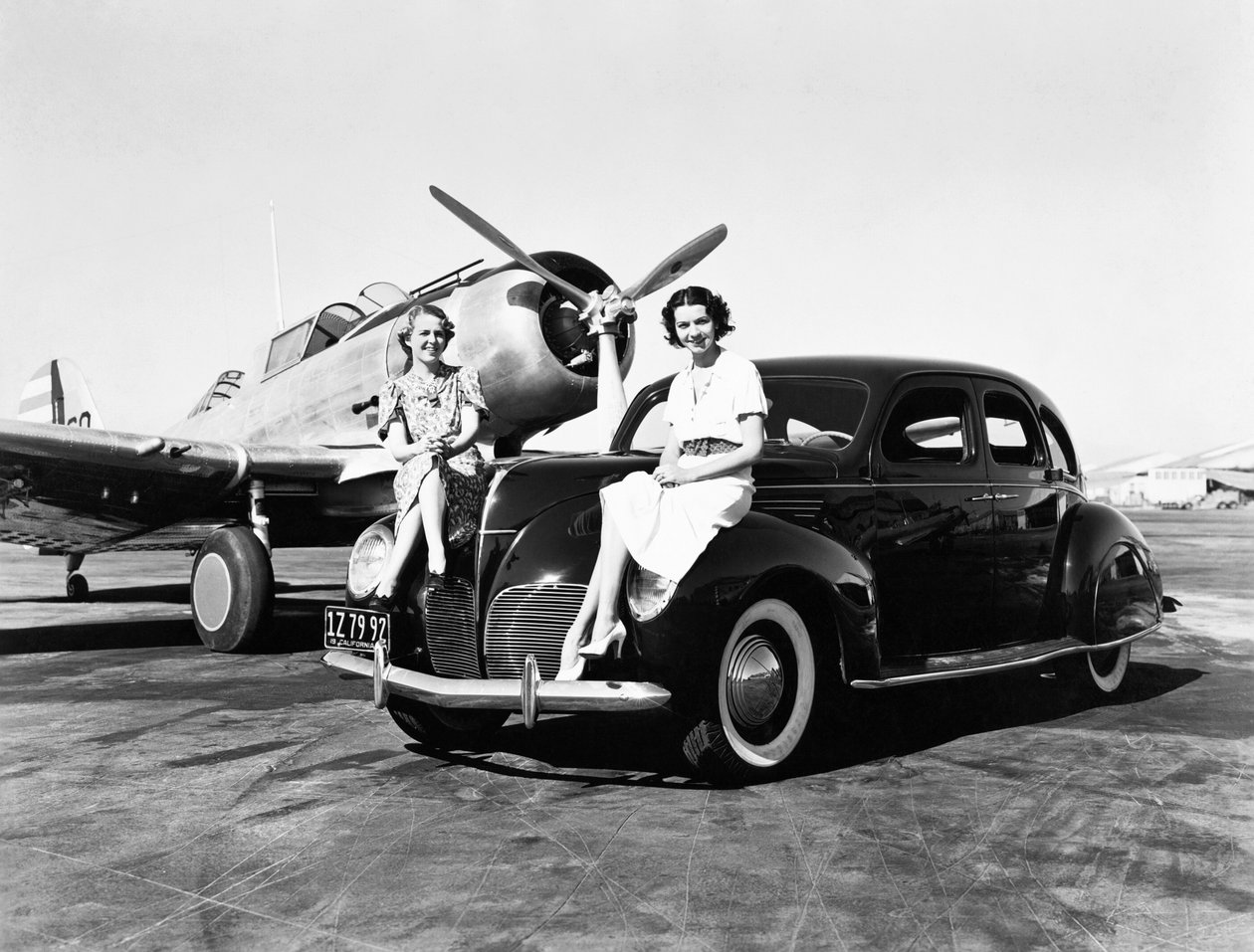 California: 1939 Due donne sedute sui parafanghi di una Lincoln Zephyr del 1939 in un aeroporto con un aereo monomotore parcheggiato dietro di loro (foto b/n) da Unbekannt