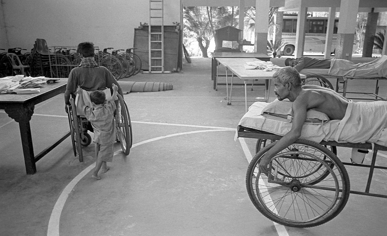 Un uomo con disabilità fisica su una sedia a rotelle viene trasportato da suo figlio; un altro uomo con la stessa condizione lo osserva al Centro per la Riabilitazione dei Paralizzati, CRP a Savar, Dhaka, Bangladesh. 2005. da Unbekannt Unbekannt