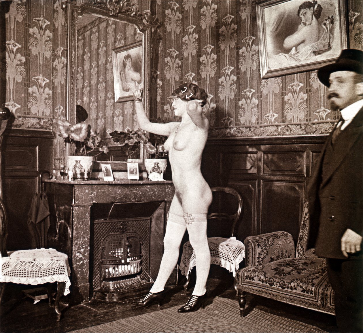 Fotografia enigmatica che mostra una giovane donna completamente nuda (tranne che per le scarpe e le calze) che si pettina davanti a uno specchio in presenza di un uomo vestito e con un cappello. Si potrebbe pensare che fosse una foto scattata in un bordel da Unbekannter Künstler
