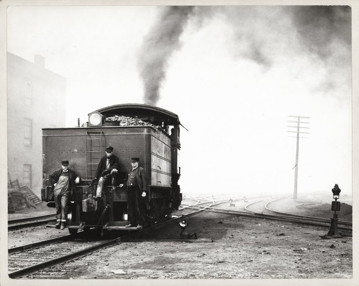 Tre uomini sul retro di una locomotiva in un deposito ferroviario non identificato da Unknown