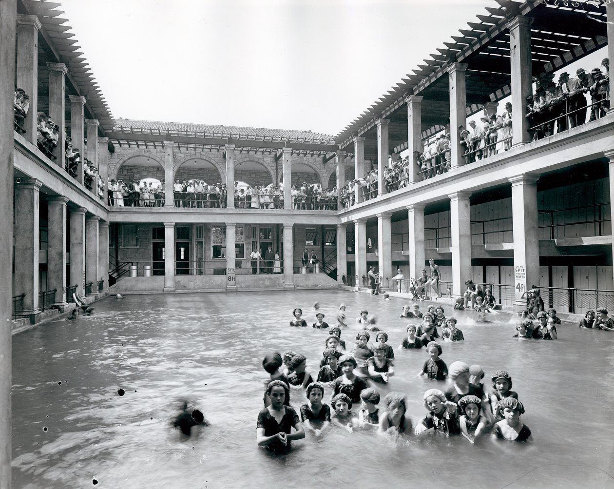Fotografia orizzontale in bianco e nero di una piscina con balcone a due piani sostenuto da colonne. Gli spettatori osservano i nuotatori dal balcone del secondo livello da William G. Swekosky