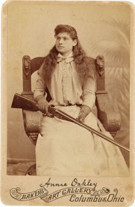 Annie Oakley: Annie Oakley. Qui la leggendaria tiratrice si siede piuttosto timidamente su una massiccia sedia in stile rinascimentale, con un fucile appoggiato sulle ginocchia. I suoi delicati tratti femminili e i capelli fluenti smentiscono la sua immagi da American Photographer