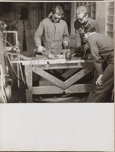 [Tre uomini con gli occhiali saldano un pezzo di metallo su un tavolo.] da Arnold Eagle