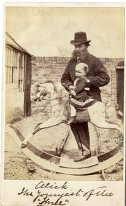 Bambina su un cavallo a dondolo in giardino, con il padre accanto da English Photographer