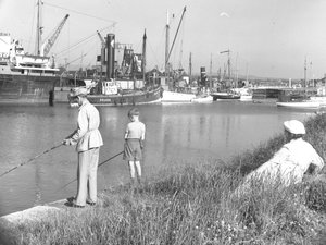 Un uomo e un ragazzo che pescano a Glasson Dock, mentre una signora naviga in mare; paesaggio marino; alle spalle, varie barche ormeggiate