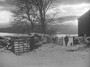 Uomo e cane che attraversano il cancello verso la telecamera nella neve con Thirlmere alle spalle da Joseph Hardman