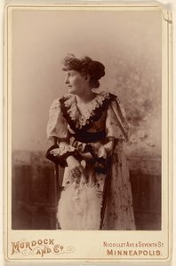 [Donna non identificata di profilo, appoggiata a una sedia, con in mano un ventaglio di piume].