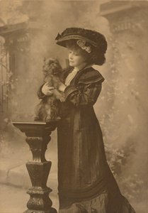 [Sarah Bernhardt in piedi in uno studio, tiene in mano un cagnolino nero su un piedistallo di legno intagliato e decorato]. da Theodore C. Marceau
