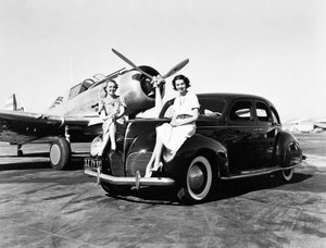 California: 1939 Due donne sedute sui parafanghi di una Lincoln Zephyr del 1939 in un aeroporto con un aereo monomotore parcheggiato dietro di loro (foto b/n)