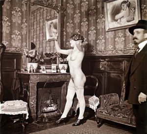 Fotografia enigmatica che mostra una giovane donna completamente nuda (tranne che per le scarpe e le calze) che si pettina davanti a uno specchio in presenza di un uomo vestito e con un cappello. Si potrebbe pensare che fosse una foto scattata in un bordel