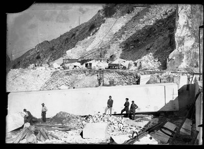 Tre uomini osservano il lavoro di diversi escavatori nelle Alpi Apuane. Dietro di loro un enorme blocco di marmo già squadrato e pronto per essere trasportato a valle da A. Corsini