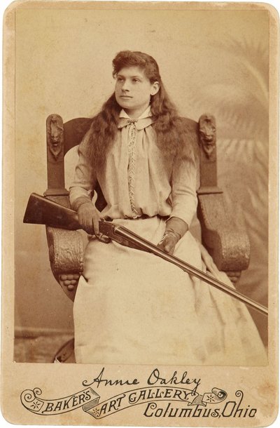 Annie Oakley: Annie Oakley. Qui la leggendaria tiratrice si siede piuttosto timidamente su una massiccia sedia in stile rinascimentale, con un fucile appoggiato sulle ginocchia. I suoi delicati tratti femminili e i capelli fluenti smentiscono la sua immagi da American Photographer