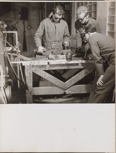 [Tre uomini con gli occhiali saldano un pezzo di metallo su un tavolo.] da Arnold Eagle