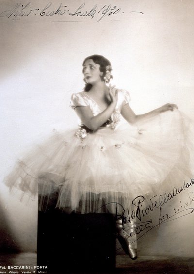 Ritratto della ballerina classica Rosa Piovella Ansaldo, in tutù e scarpette da ballo. È seduta, frontalmente, con il viso girato di lato. Rose di stoffa decorano il suo abito e i capelli accuratamente acconciati (Milano, Teatro alla Scala) da Baccarini and Porta