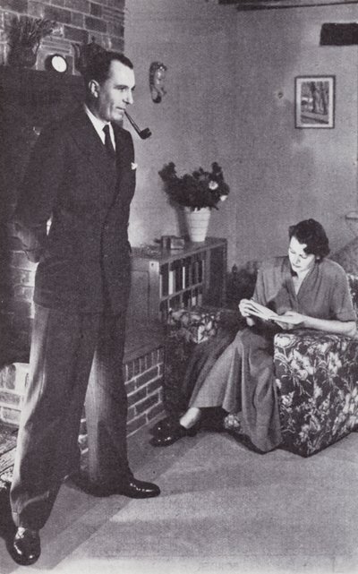 La moglie legge, mentre il marito si scalda la schiena contro il fuoco, c.1950 da English Photographer