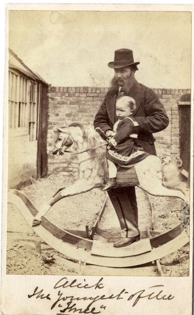 Bambina su un cavallo a dondolo in giardino, con il padre accanto da English Photographer