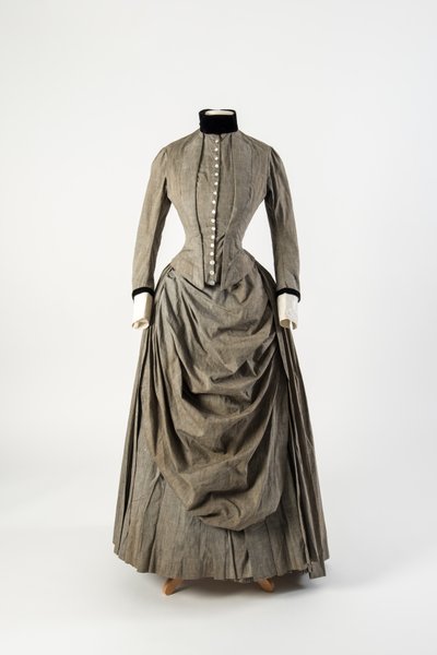 Abito di cotone grigio con grembiule drappeggiato sul davanti e rifiniture in velluto nero al colletto e ai polsini, fine 1880 da English School