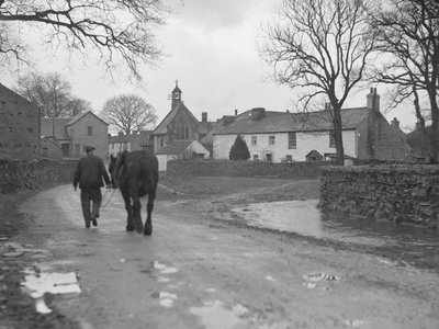 Uomo che conduce il cavallo lungo la strada, lontano dalla telecamera, verso gli edifici del villaggio e la chiesa; architettura; religioso; ecclesiastico; da Joseph Hardman