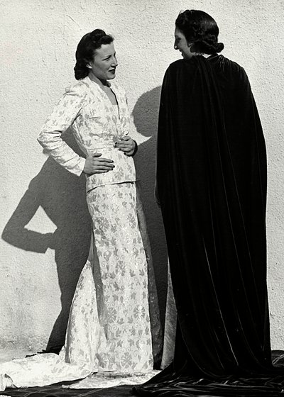 Due modelle presentano creazioni della casa di moda italiana "Dragoni". Una modella indossa un abito in tessuto operato composto da giacca e lunga gonna con strascico. L