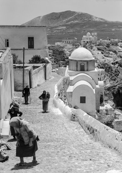 Persone di un villaggio greco camminano su una strada di ciottoli in un villaggio greco con edifici imbiancati da Maynard Owen Williams