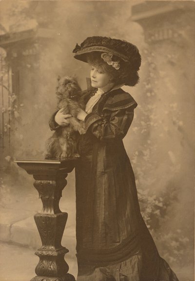 [Sarah Bernhardt in piedi in uno studio, tiene in mano un cagnolino nero su un piedistallo di legno intagliato e decorato]. da Theodore C. Marceau
