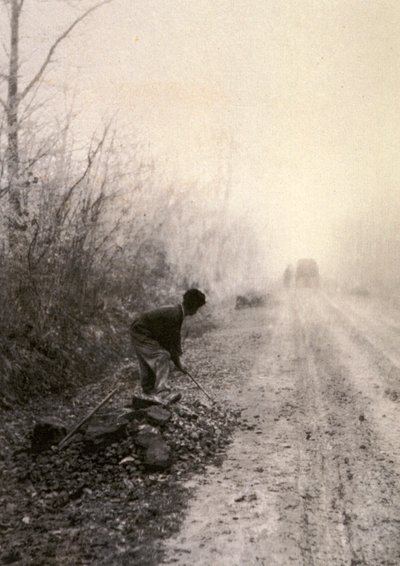 Una strada di campagna avvolta nella nebbia mattutina. In primo piano un uomo spacca pietre da Unbekannter Künstler