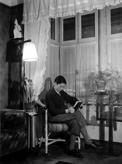 Un adolescente seduto in un interno borghese legge un libro, indossa pantaloni corti e calze alte. 1915. Foto storica, donna che legge, ca. 1915 da Unbekannter Künstler