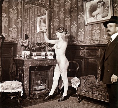 Fotografia enigmatica che mostra una giovane donna completamente nuda (tranne che per le scarpe e le calze) che si pettina davanti a uno specchio in presenza di un uomo vestito e con un cappello. Si potrebbe pensare che fosse una foto scattata in un bordel da Unbekannter Künstler