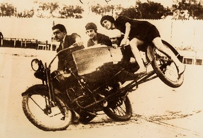 Un uomo si esibisce guidando una motocicletta con un sidecar che trasporta tre giovani donne. Il sidecar si solleva da terra mentre prendono una curva. da Unknown photographer