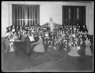 Ritratto di gruppo di una classe di coro di ragazze riunite attorno a un pianoforte con spartiti, probabilmente presso l