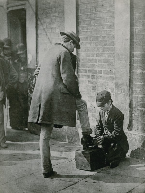 Un giovane lustrascarpe che lucida le scarpe di un uomo per strada da English Photographer