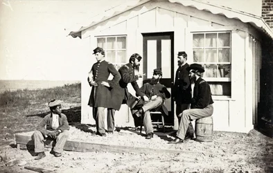 Cinque soldati della Guerra Civile riuniti su un portico di terra fuori casa, giovane afroamericano seduto vicino a loro, 1861-65