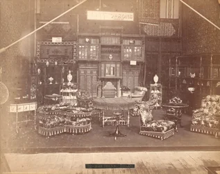 Esposizione di S. Howard & Sons -- Edificio principale, Collezione di mobili, inclusi sedie imbottite, tavoli, un camino, armadi e scaffali. Sulle pareti ci sono tappeti, e su tavoli e scaffali ci sono vasi