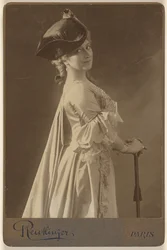 [Donna non identificata che indossa un cappello scuro, in piedi, con le mani appoggiate su un bastone da passeggio].