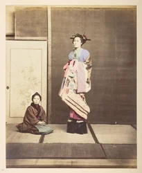 [?Geisha,] Ritratto in studio a figura intera di una giovane donna con scarpe elevate, il suo giovane assistente inginocchiato sul pavimento accanto a lei