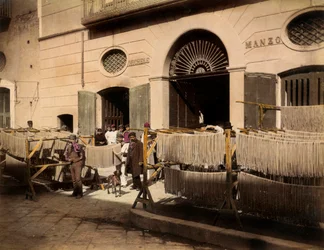 File di essiccatoi di maccheroni davanti a una fabbrica a Torre Annunziata. La scena è animata dagli operai del pastificio: alcuni sono in posa, in primo piano, vicino agli essiccatoi, altri sono sulla soglia degli ingressi della fabbrica