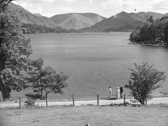 Un uomo, una donna e forse la loro figlia passeggiano lungo il bordo di un lago, sullo sfondo le colline.