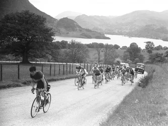 Gruppo di ciclisti che sale su una strada seguito da automobili, sullo sfondo il lago e le montagne