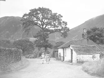 Due giovani ragazze che si tengono per mano tenendo rametti di foxglove camminano lungo la strada passando davanti a un cottage verso l