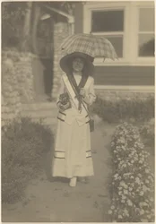 [Donna in piedi nel giardino davanti casa con una macchina fotografica e un ombrello a quadri]